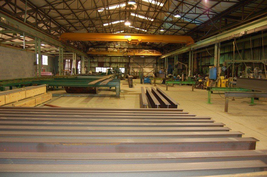 Warehouse interior with steel beams. Overhead crane, worktables, and equipment present.
