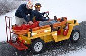 Yellow rescue vehicle carrying a patient on a stretcher, with two people assisting on a snow-covered road.