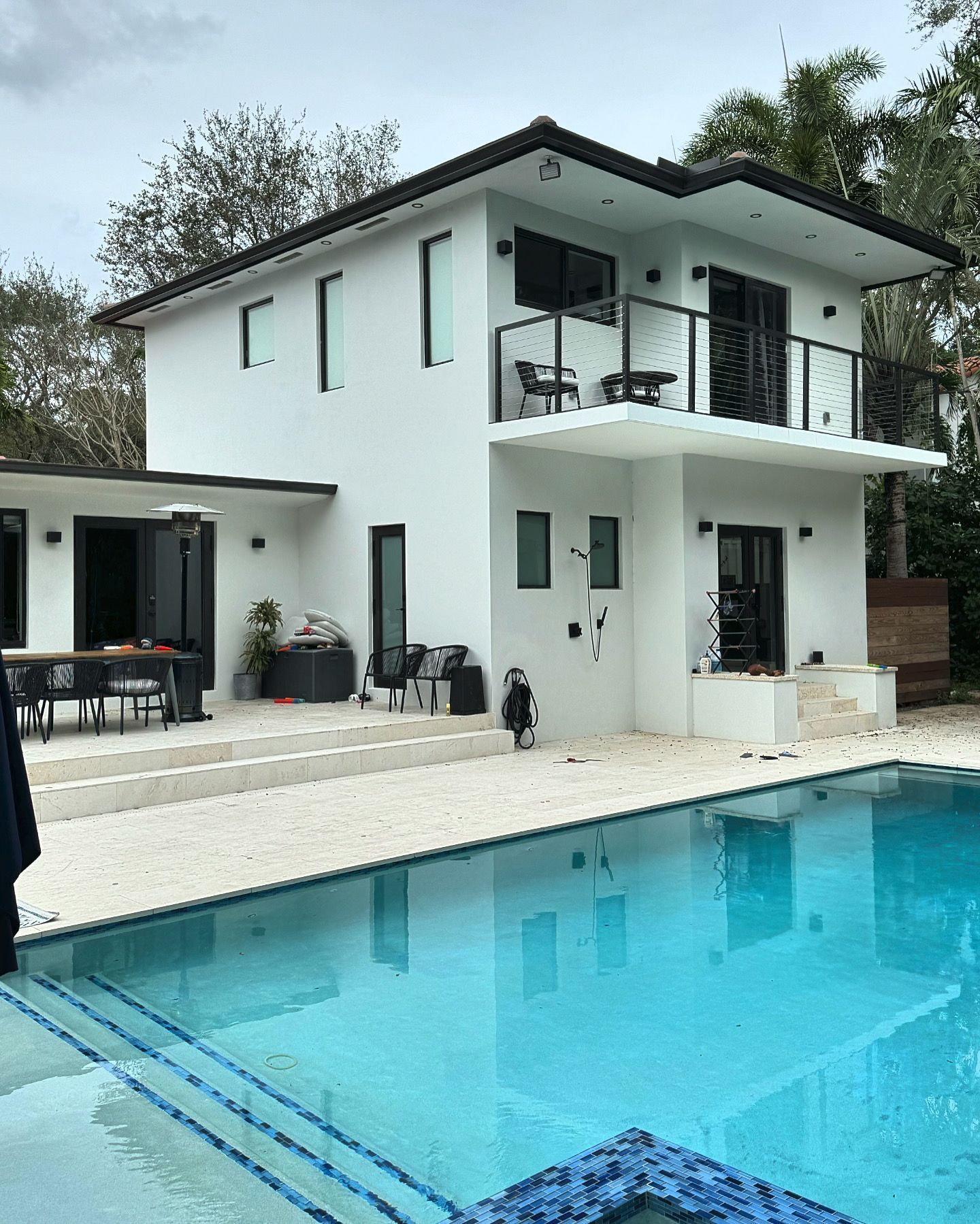 Two-story white house with balcony overlooking a turquoise pool. Outdoor patio with table and chairs.