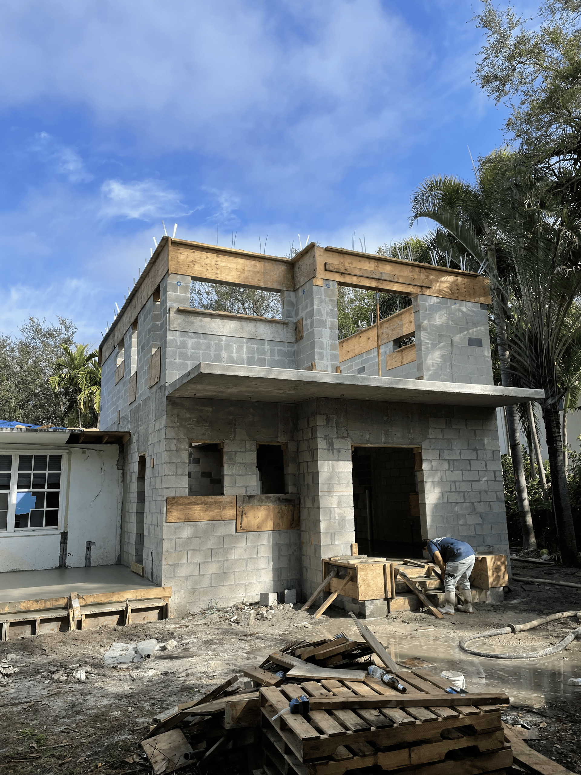 Two-story concrete block house under construction; a worker stands in front, debris in foreground; cloudy sky.