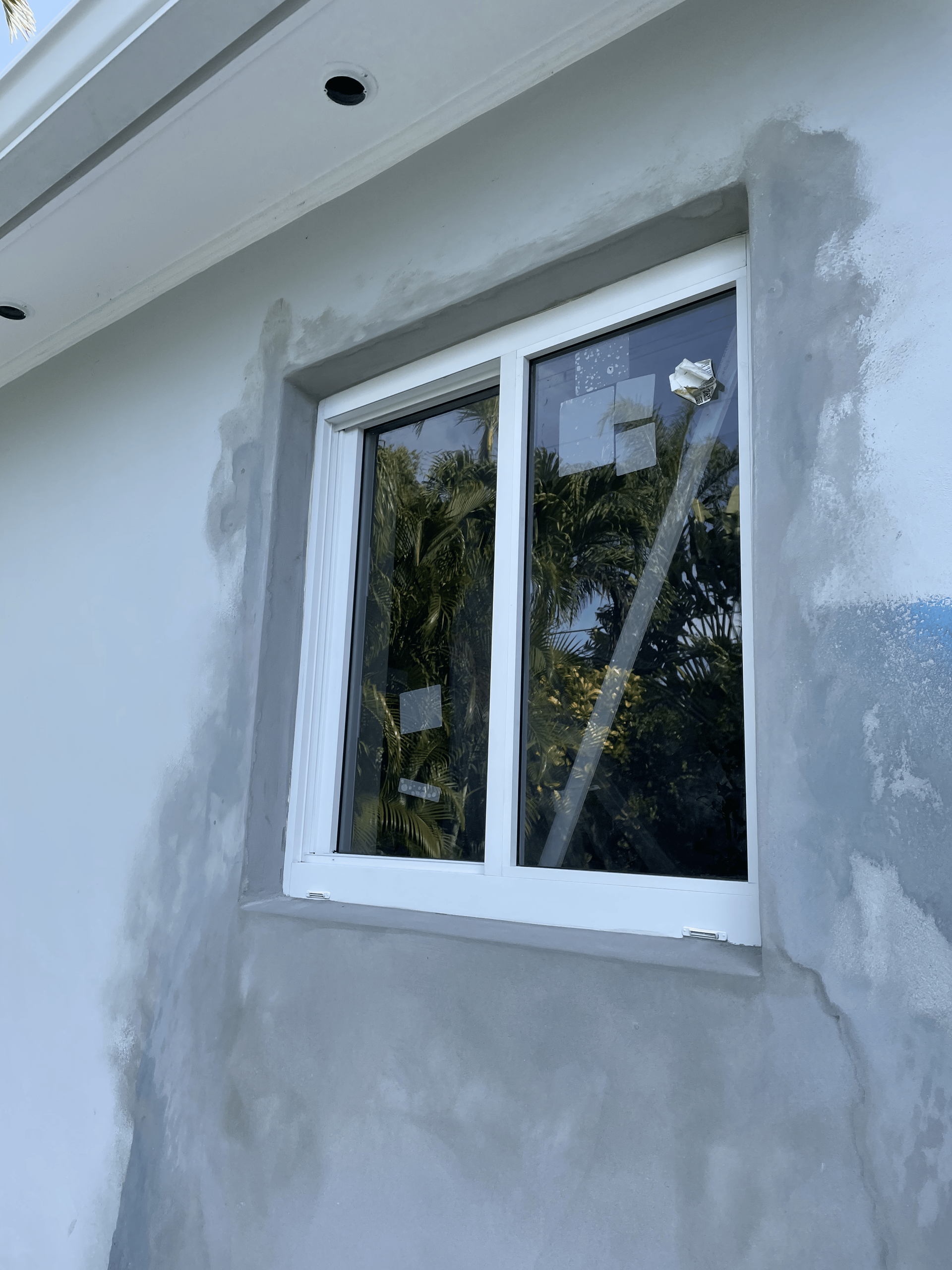 White-framed window on a light gray wall with visible plaster patches, looking out onto trees.