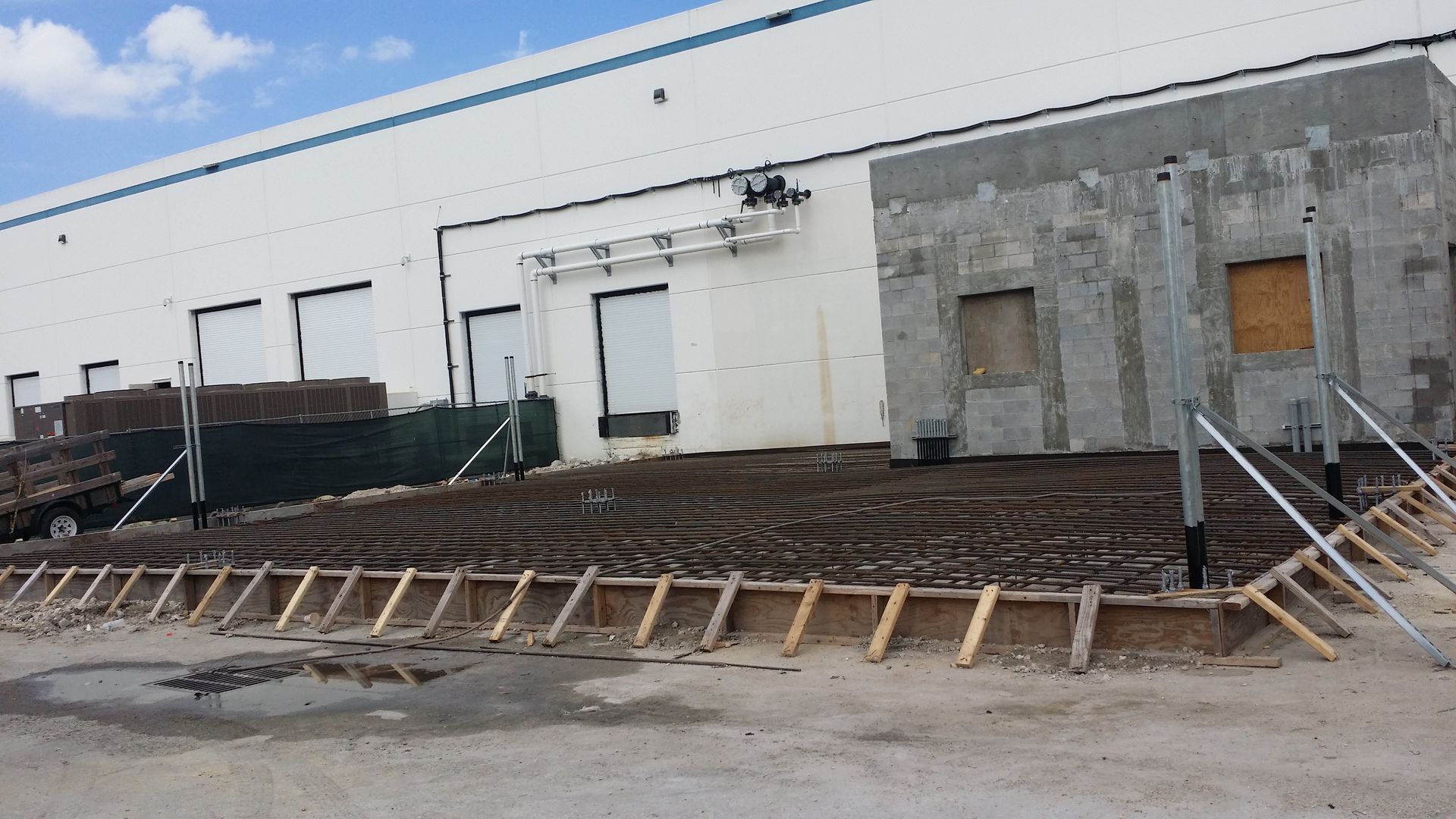Construction site next to a white industrial building; wooden forms and dirt visible.