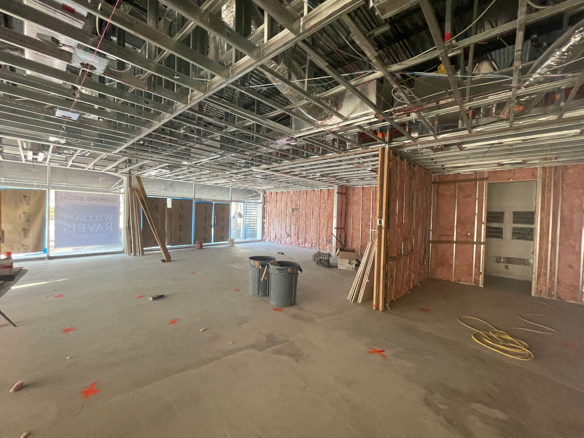 Interior construction site: exposed metal ceiling beams, insulated walls, concrete floor, windows, construction materials.