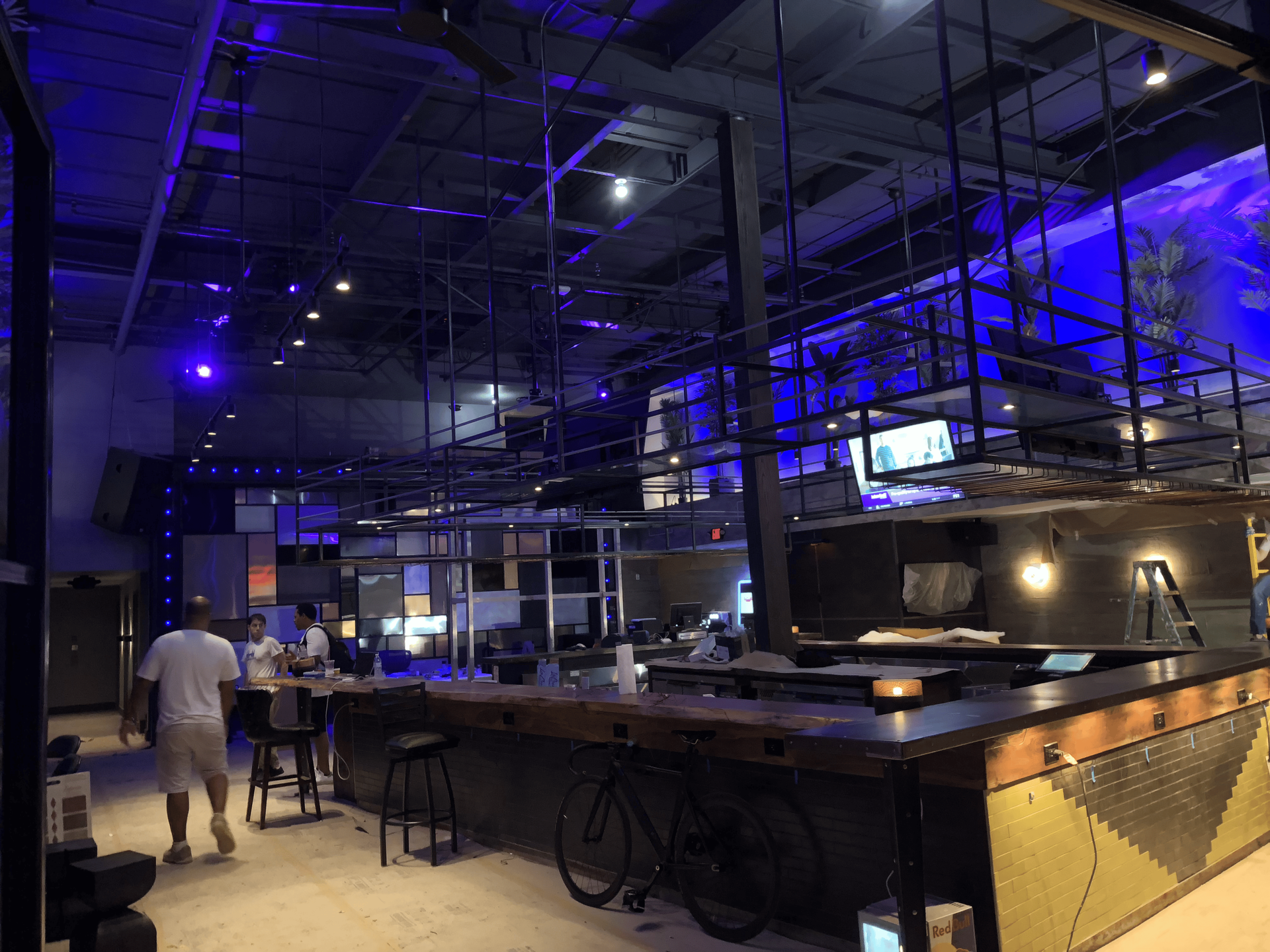 Interior of a modern bar with a long wooden counter. People sit at the counter and tables. Blue lighting.