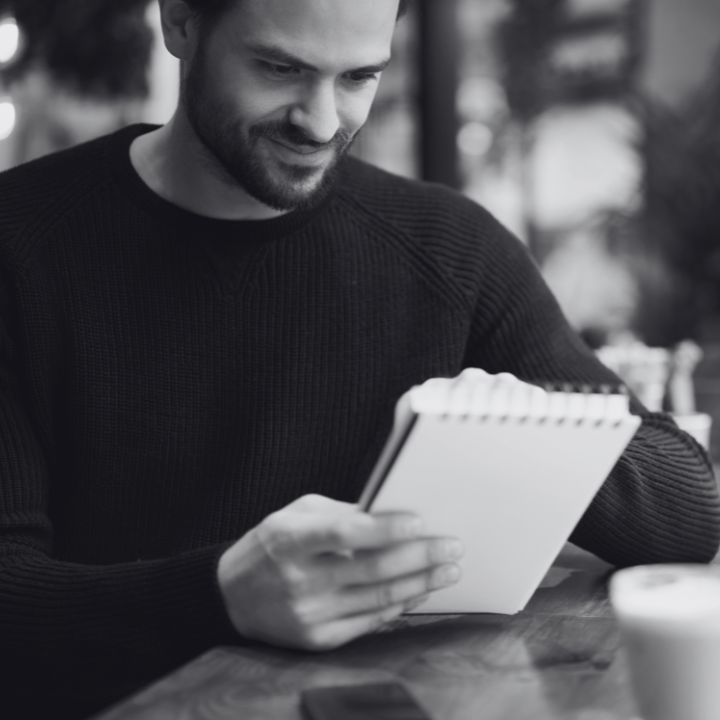 Man in black sweater, smiling, writing in a notebook at a table in a cafe. Black and white.