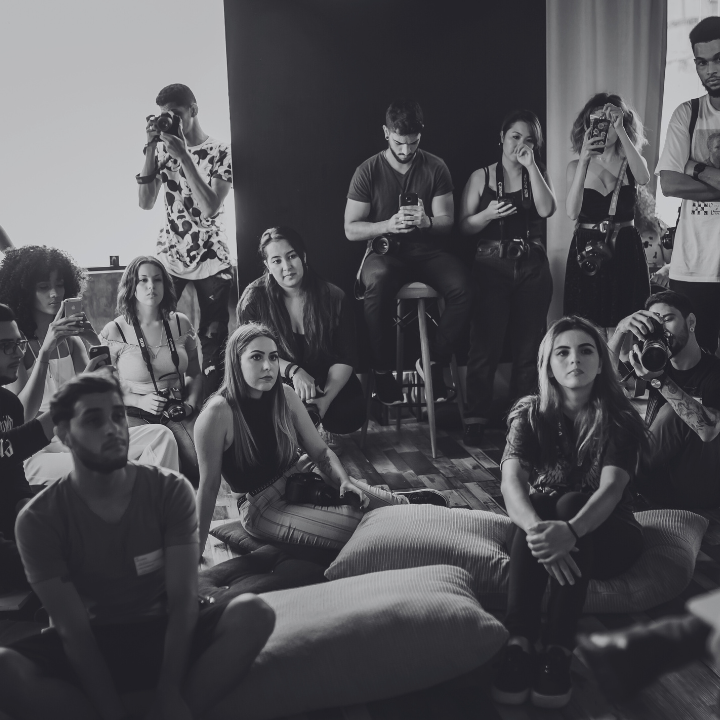 Group of people in a room; some sit on cushions, others stand, and several hold cameras. Black and white.