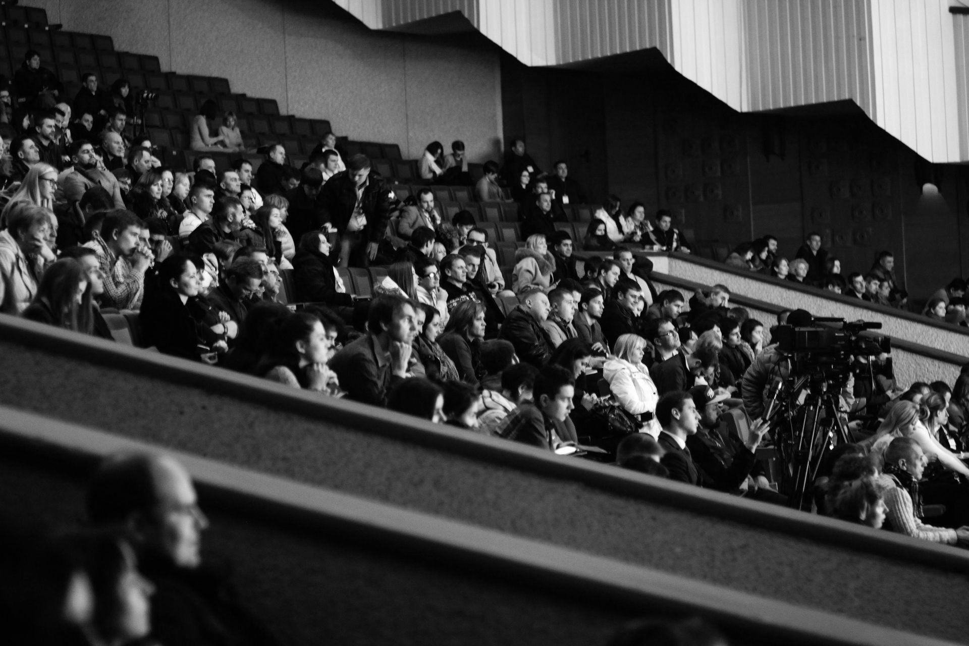 Audience seated in tiered theater seating, watching a performance. Black and white photo.