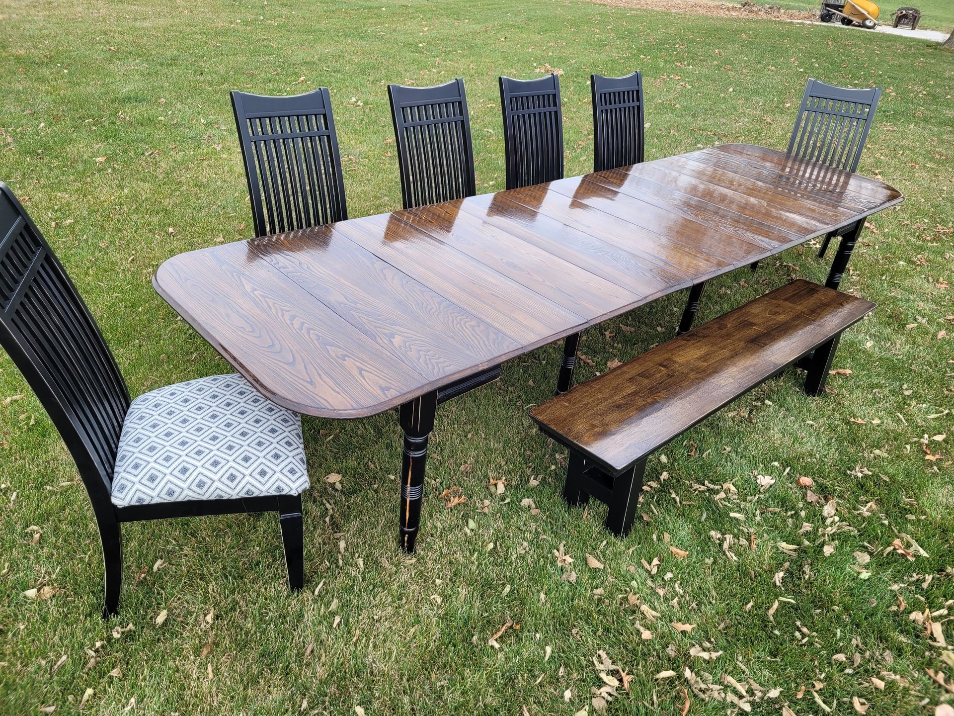 Outdoor wooden dining table with six black chairs and a matching bench on grass
