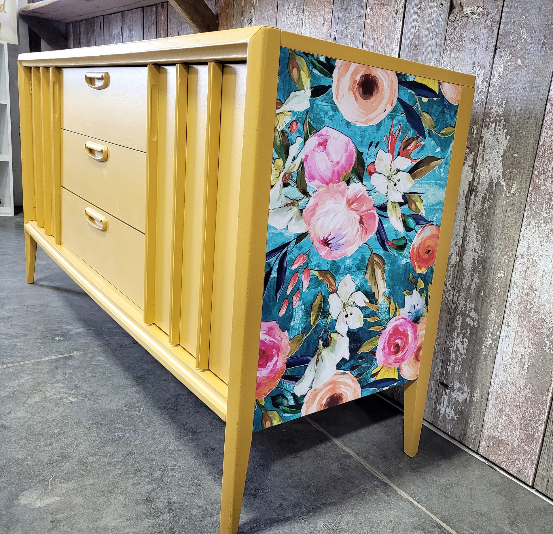 Yellow dresser with floral-patterned side panel on a concrete floor, leaning against a wall.