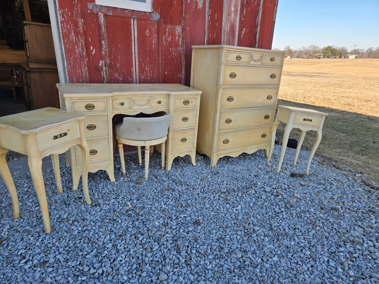 Antique cream-painted vanity, dressers, and stools arranged on gravel beside a red barn