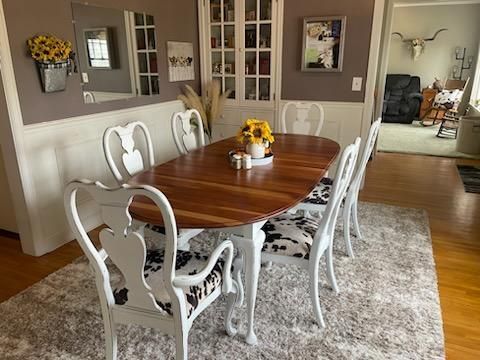 Dining room with a wood table, white chairs, floral decor, and a large rug by a hallway open to another room