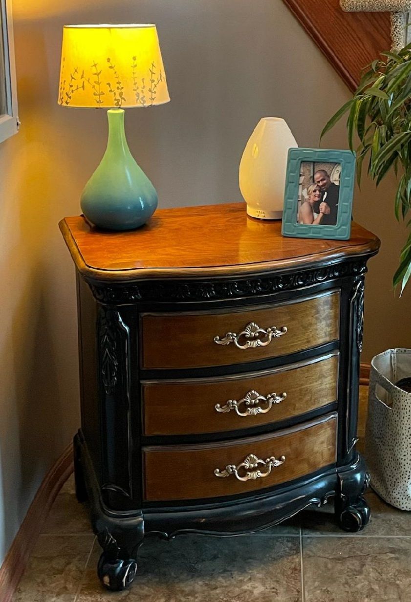 Black bedside table with three drawers, lamp, photo frame, and decorative vase on top
