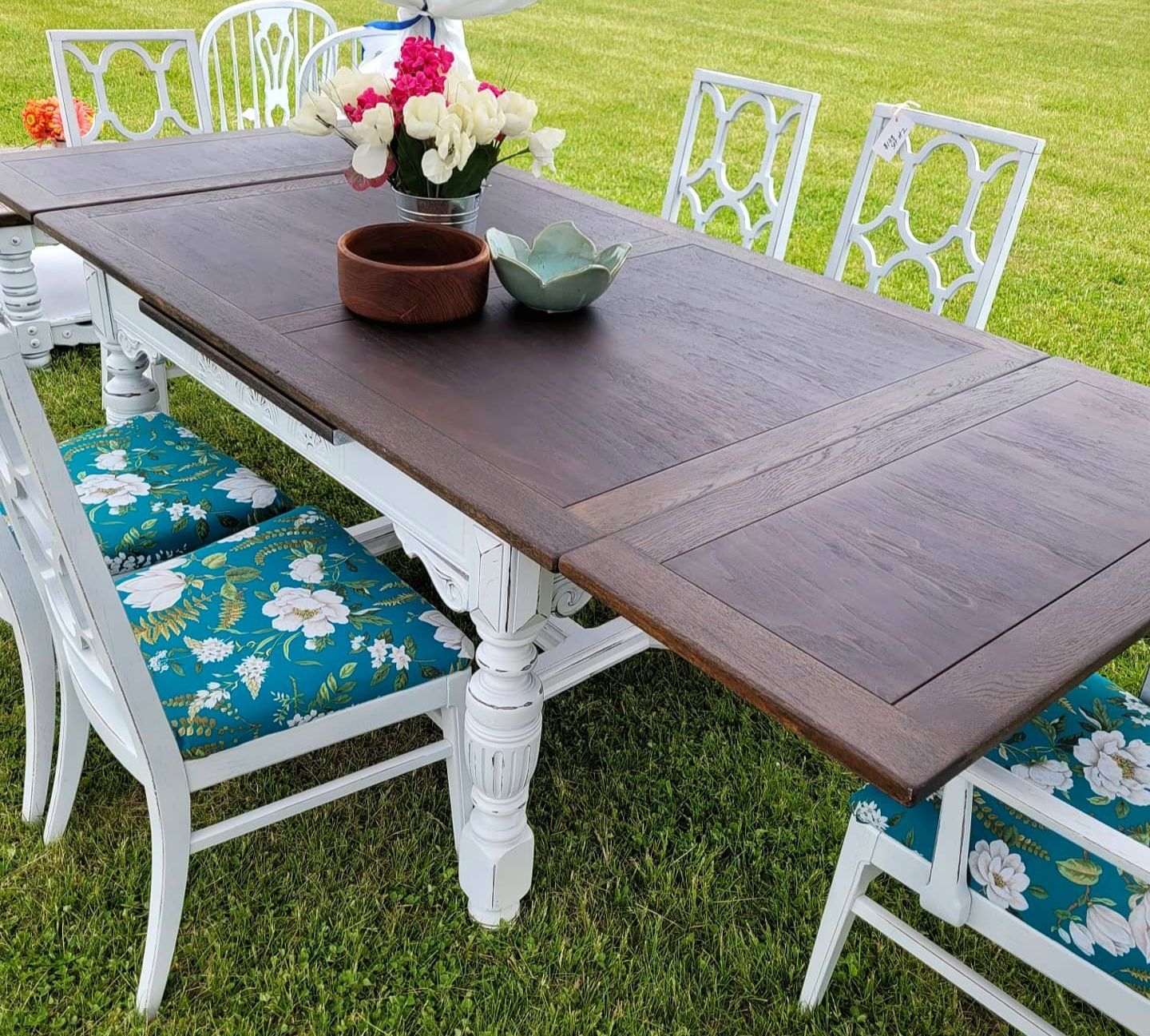 Outdoor dining table with dark wood top, white chairs, and floral cushions on grass