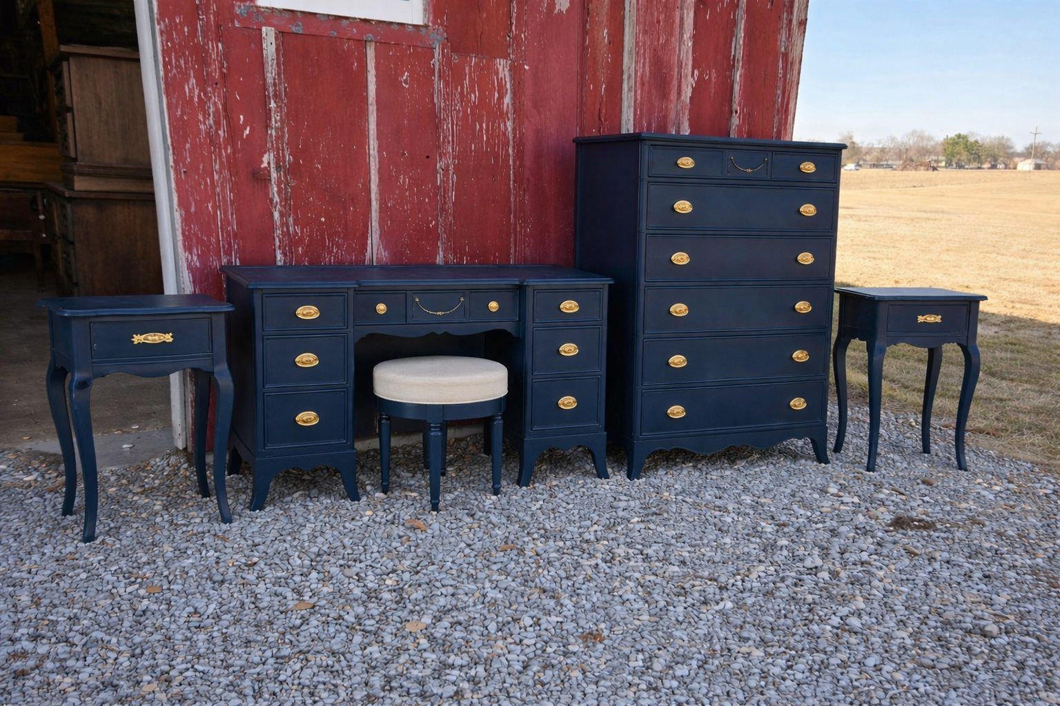 Dark blue vintage desks and side tables displayed on gravel in front of a weathered red barn.
