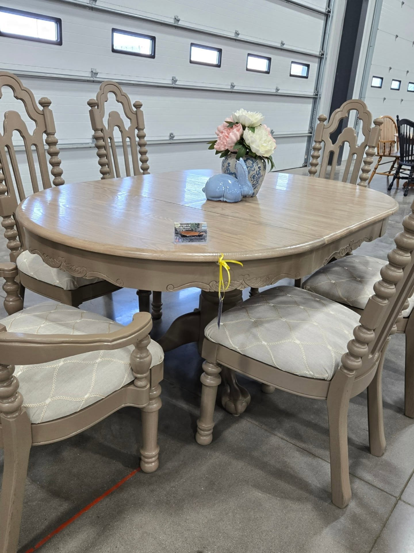 Round wooden dining table with cream chairs and a small flower centerpiece in a warehouse showroom
