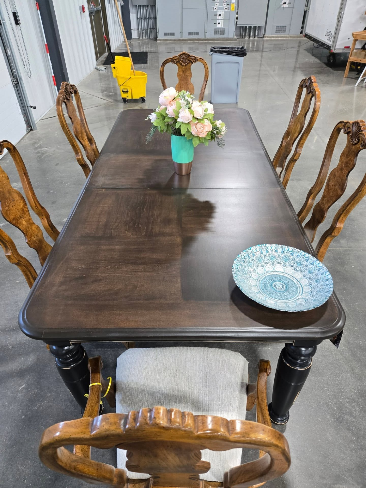 Dark wooden dining table with carved chairs, a flower vase, and a blue plate in a showroom-like space