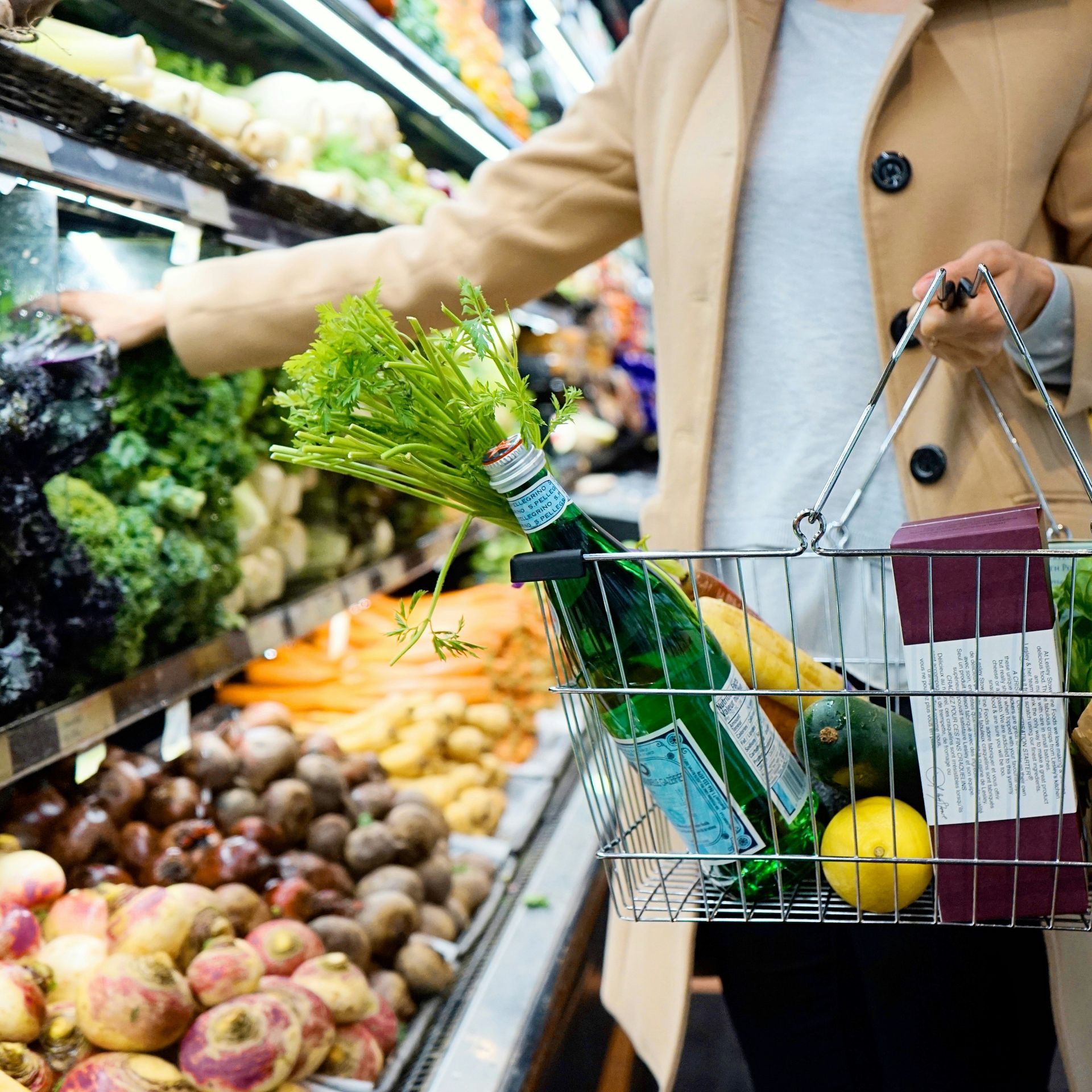 A shopper with a grocery basket selecting vegetables from the shelf.