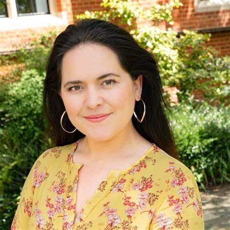 Woman with dark hair and hoop earrings, wearing a yellow floral shirt, smiles outdoors.