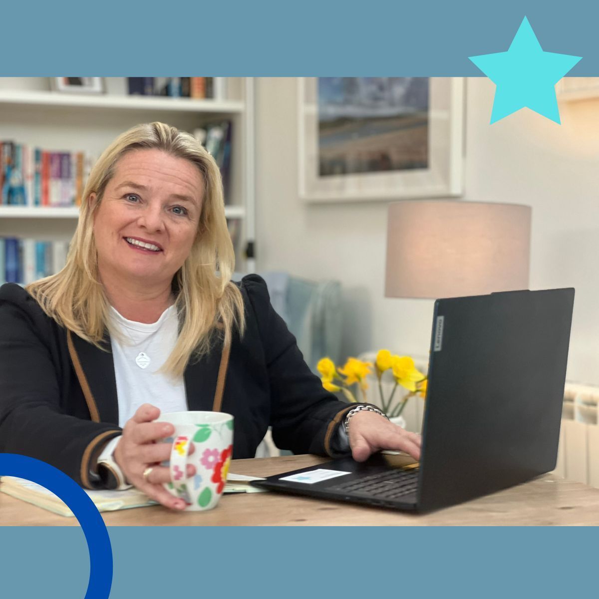 Woman with blonde hair smiles, holding a mug and using a laptop at a desk in a home office.