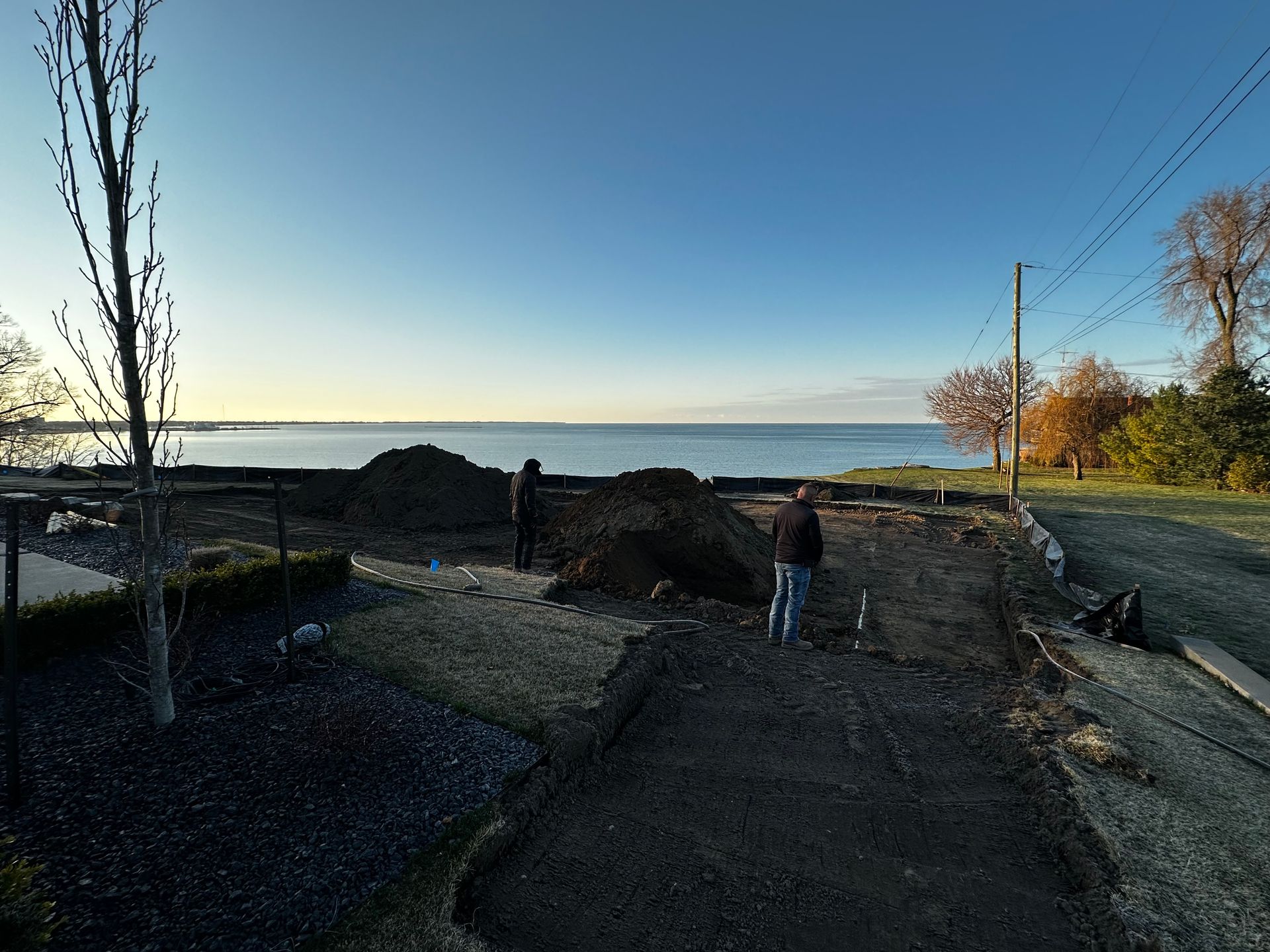 Person standing on a rocky path by the water at sunset, with trees and a clear blue sky