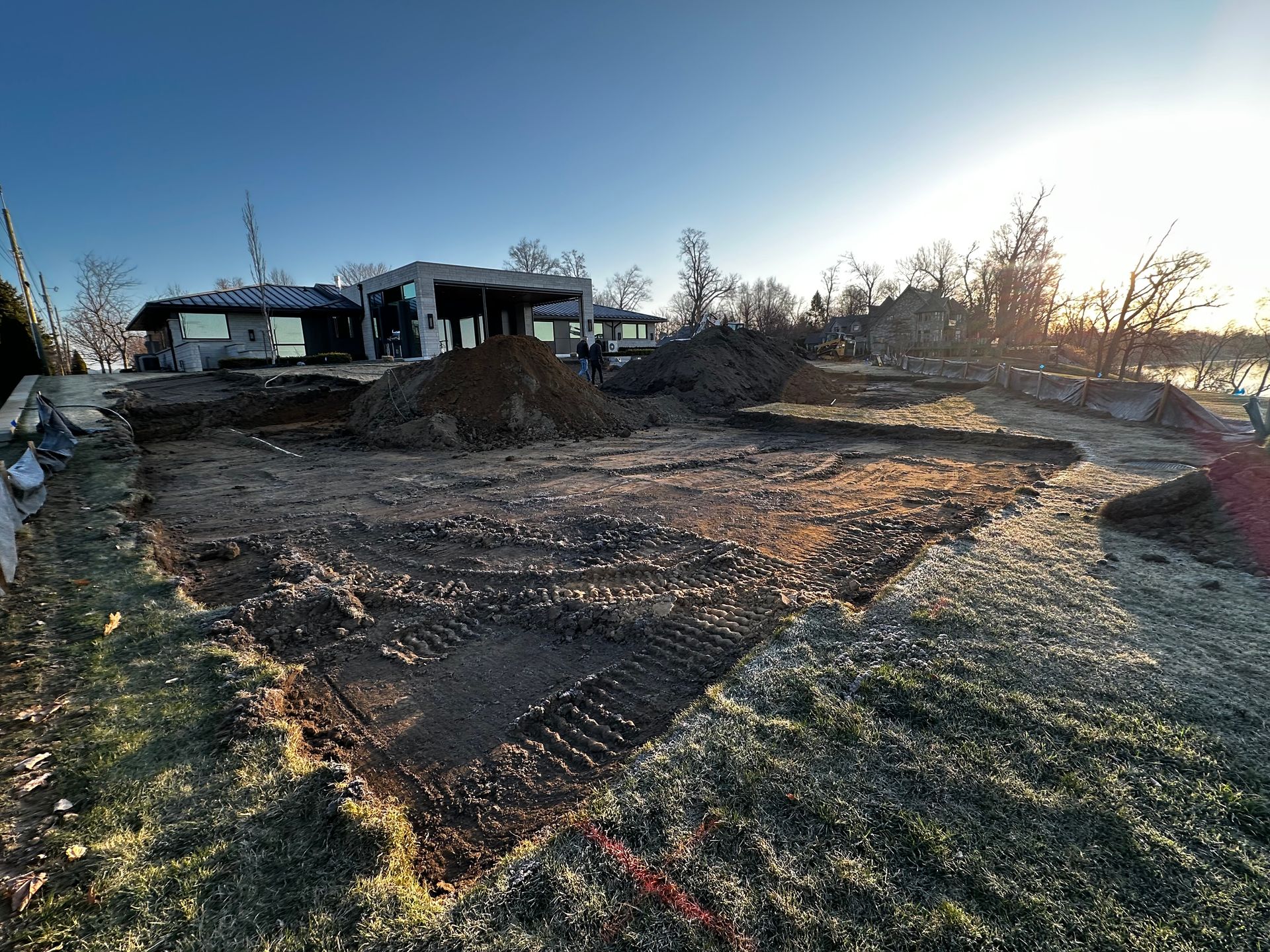 Gravel construction site at sunrise with mounds of dirt, bare trees, and houses in the background