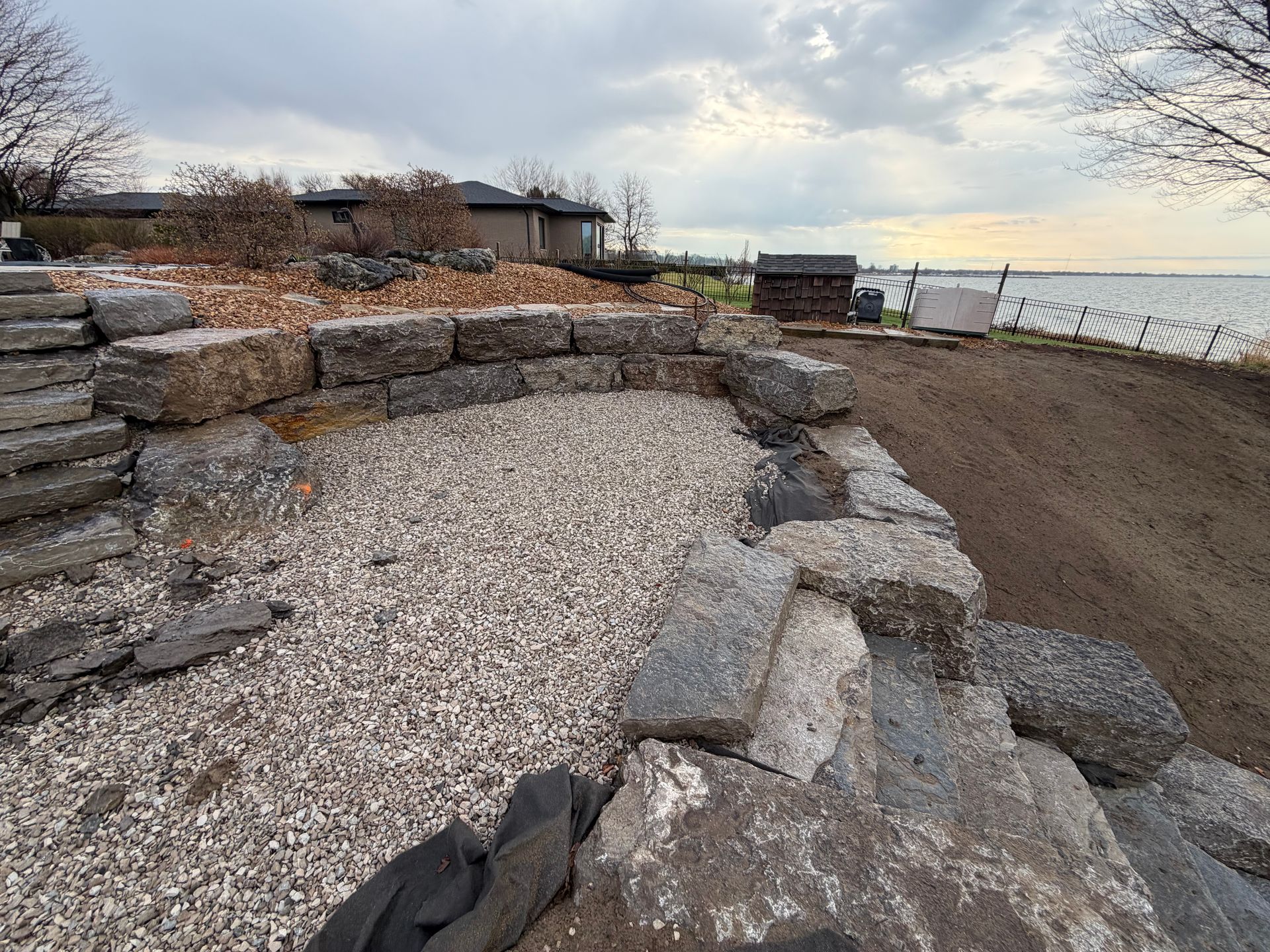 Stone retaining wall along a gravel shoreline path beside calm water under a cloudy sky
