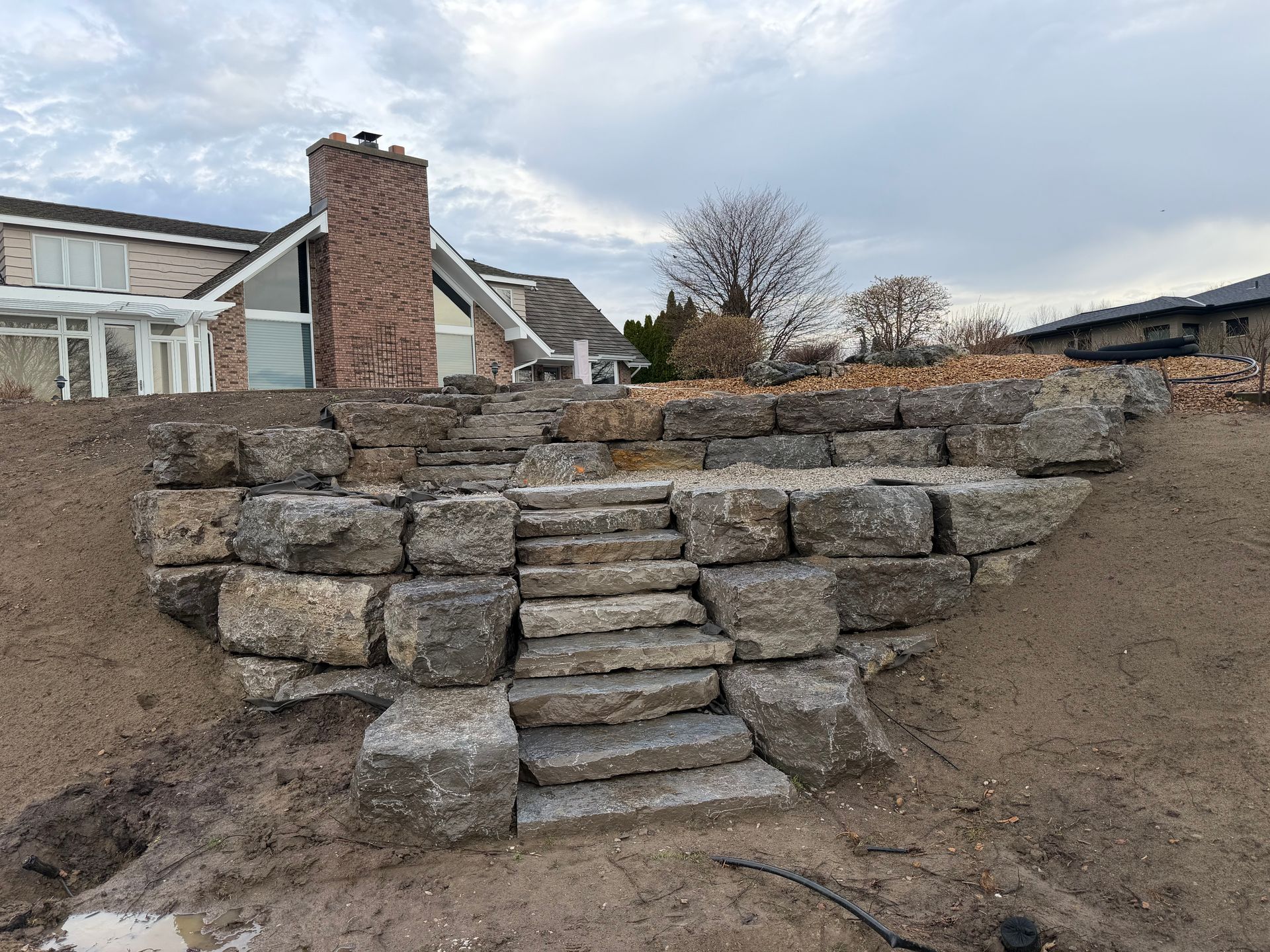 Stone steps built into a retaining wall on a sloped yard beside a house under a cloudy sky