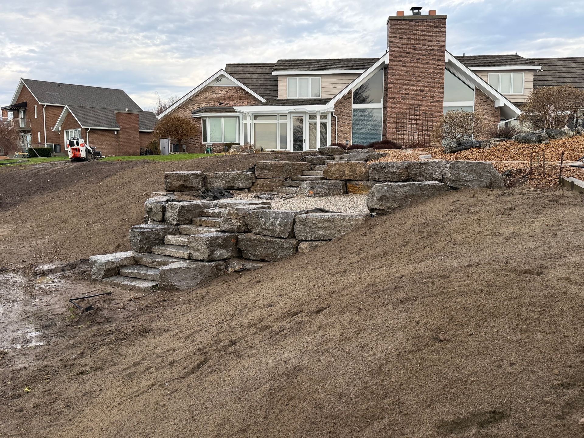 Stone steps and a retaining wall on a dirt slope in front of suburban houses.