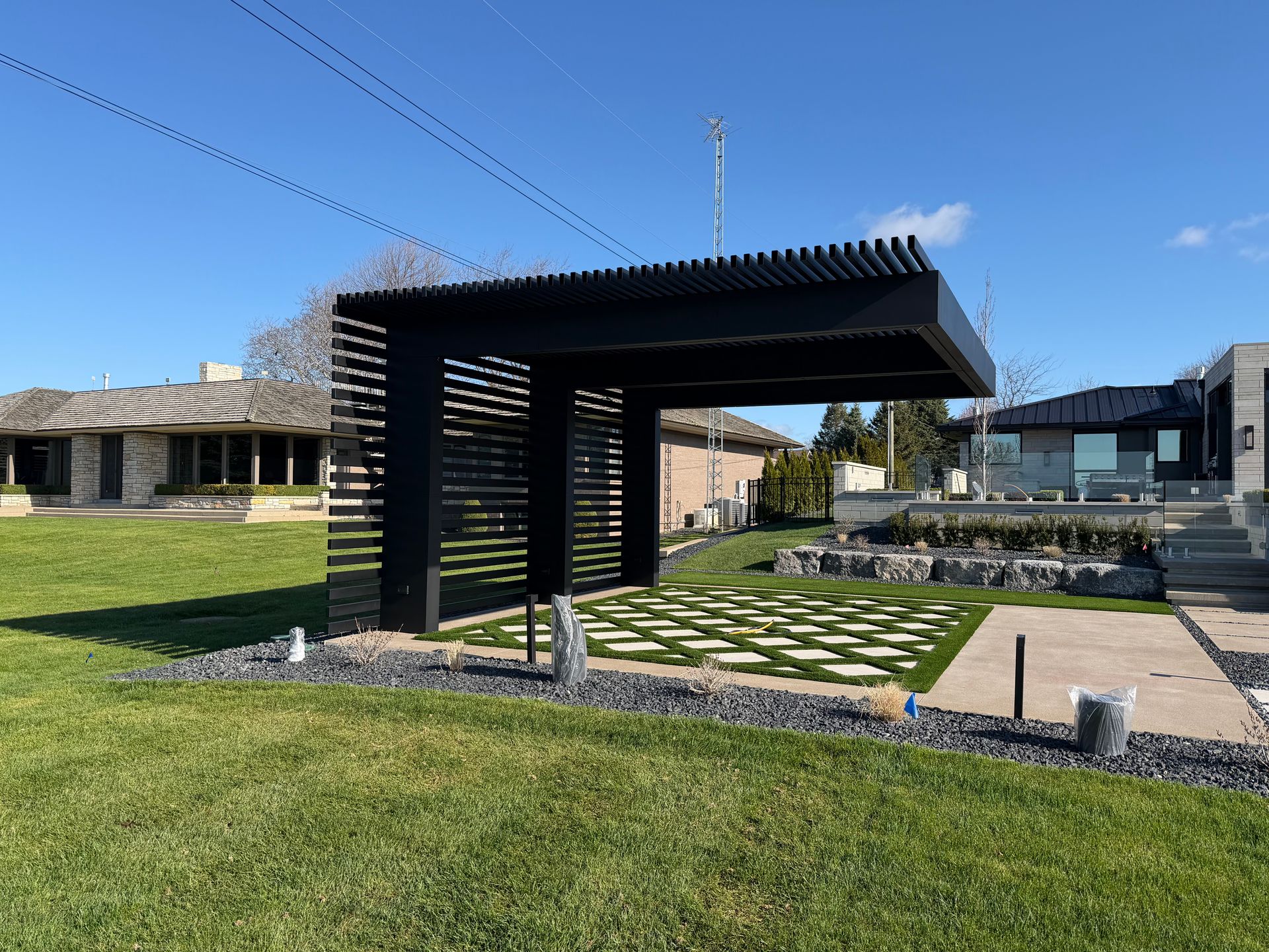Modern black pergola over a stone patio in a grassy courtyard with buildings in the background
