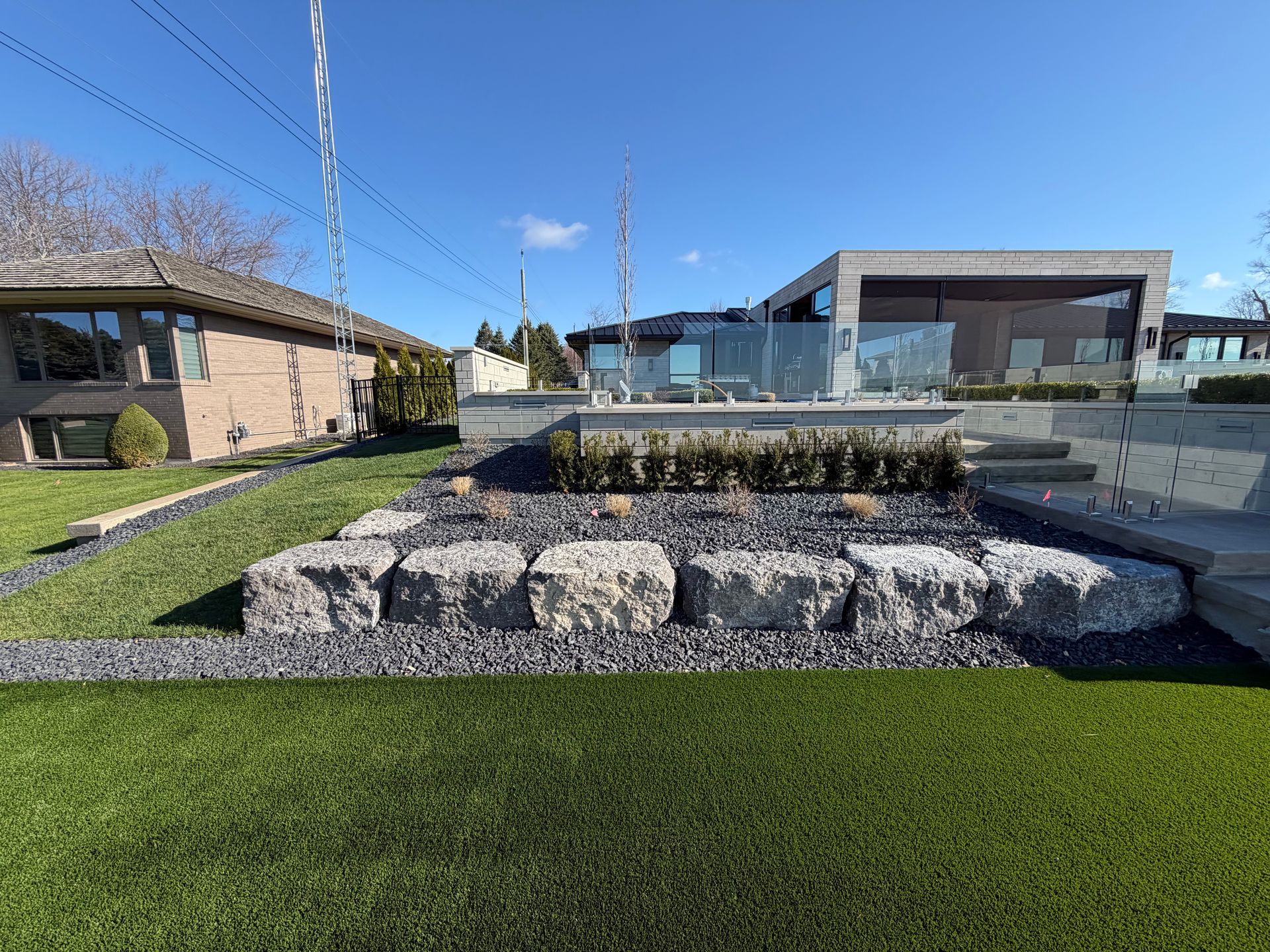 Outdoor plaza with a central fountain, stone retaining wall, green lawn, and buildings under a blue sky