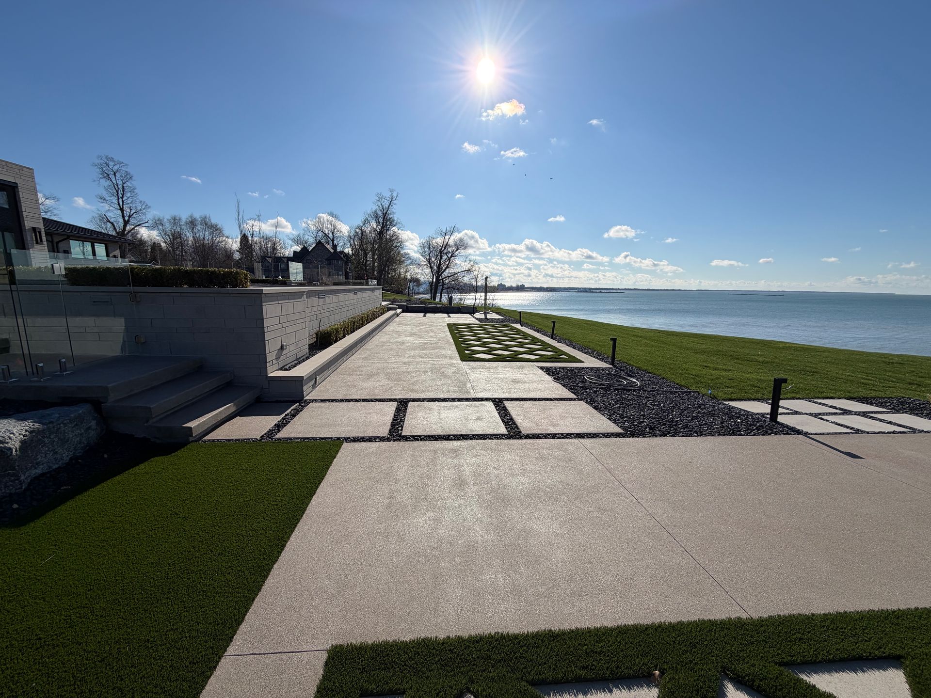 Sunny waterfront patio with stone pavers, grass, and a view of the calm sea and blue sky.