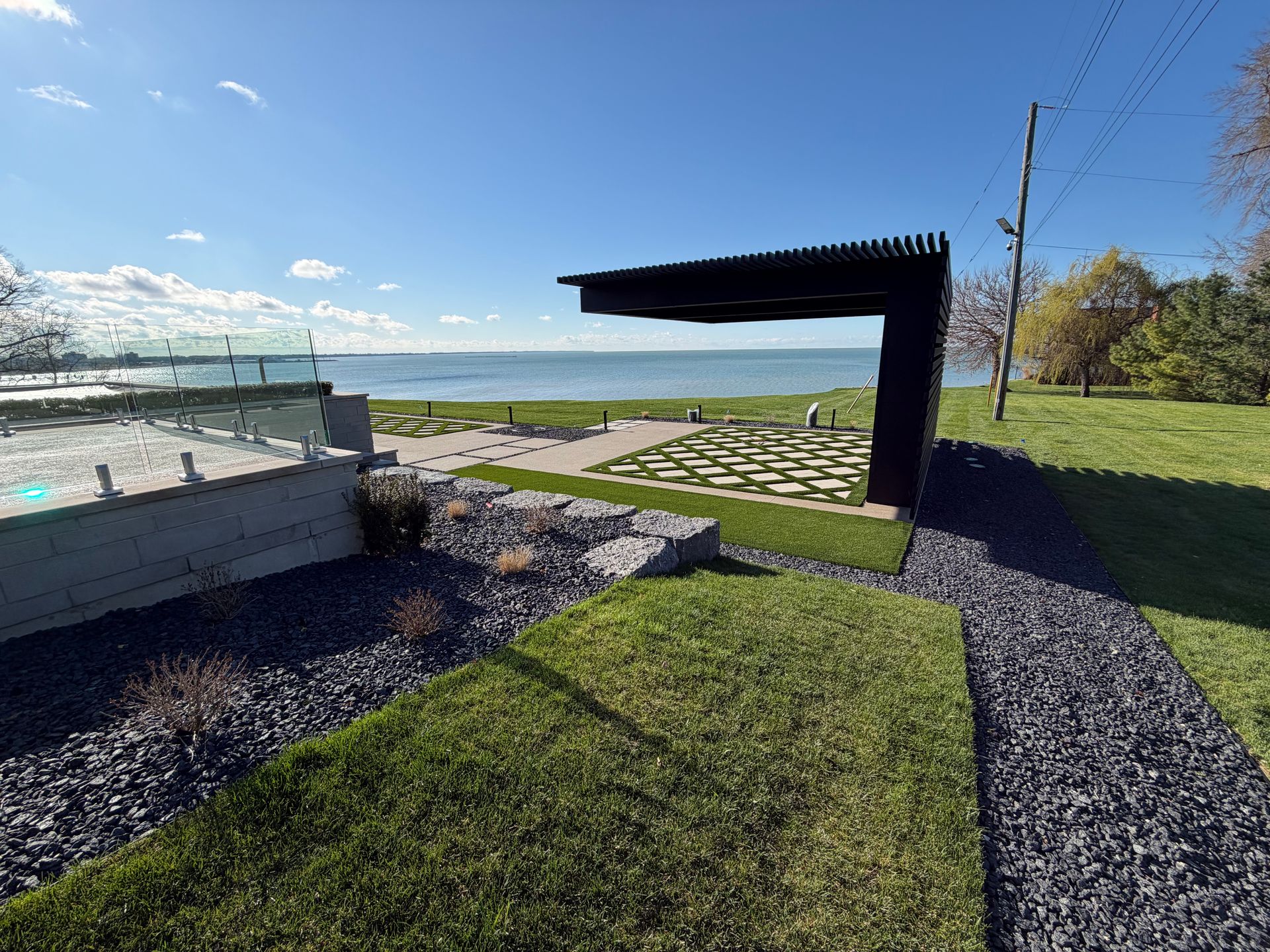 Beachfront park with a black pergola, stone path, grass lawn, and ocean view under a blue sky