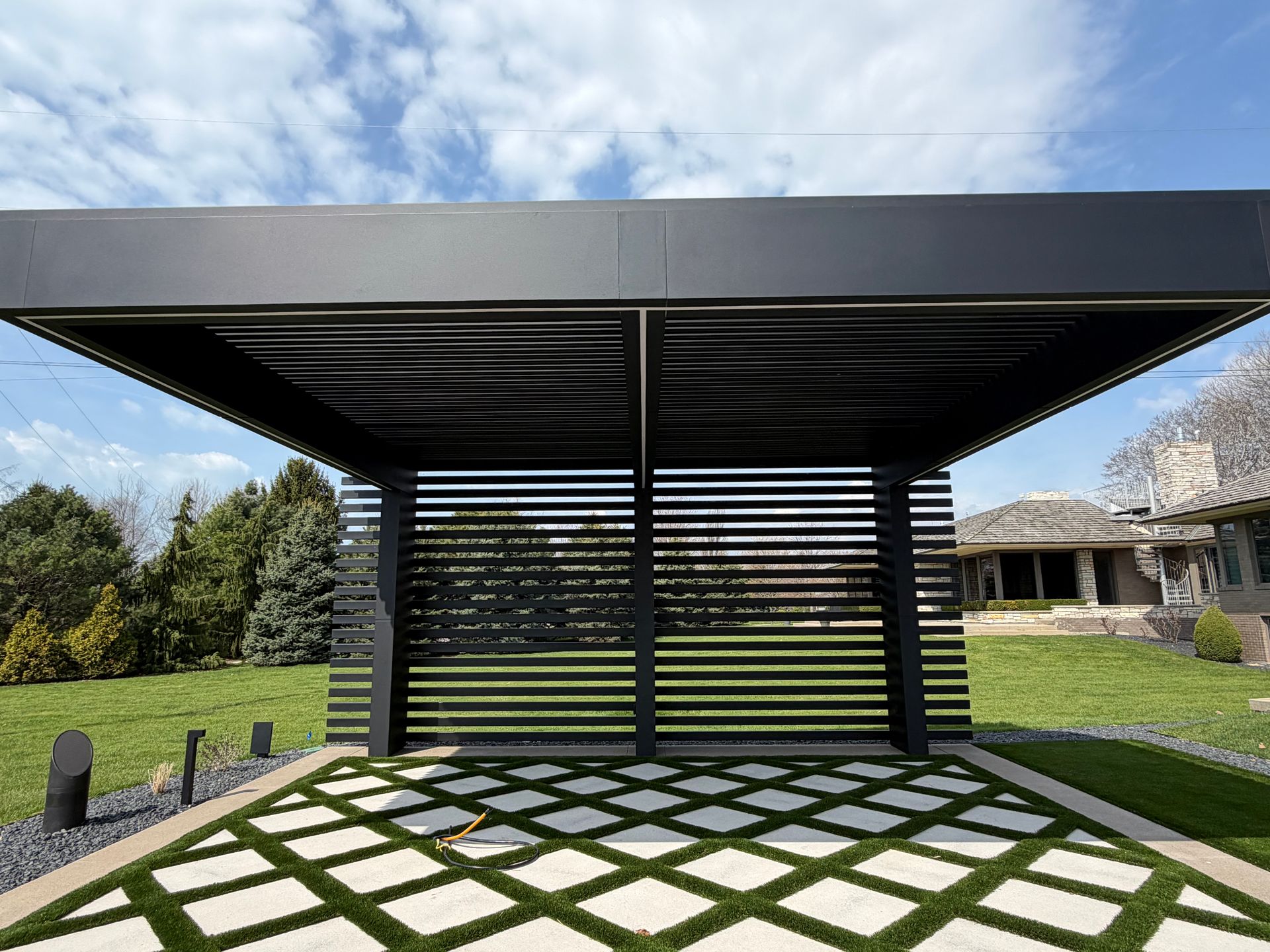 Modern black pergola with lattice walls over a patterned patio in a landscaped backyard.