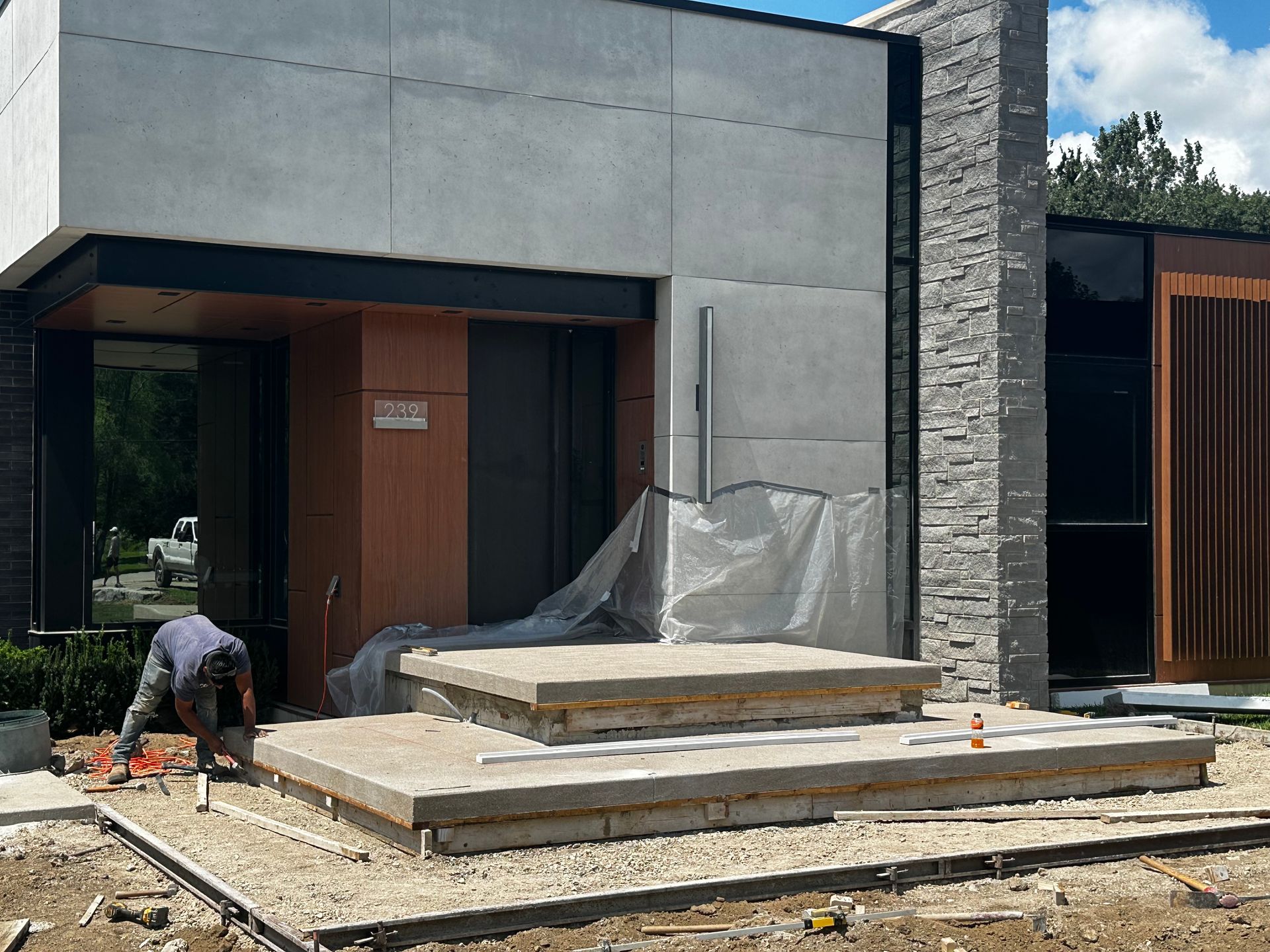 Worker at a modern house entrance, laying stone steps beside a concrete and wood facade.