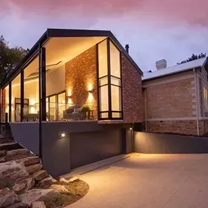 Modern glass-and-brick house glowing warmly at dusk, with a stone walkway and trees nearby