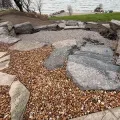 Rock garden with large stones and gravel beside a lake, with an excavator at right.