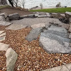 Rock garden with large stones and gravel beside a lake, with an excavator at right.