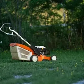 Orange push lawn mower on grass with a white collection bag, outdoors at dusk.