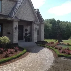 Brick house entrance with curved stone walkway, landscaped garden, and trees in the background