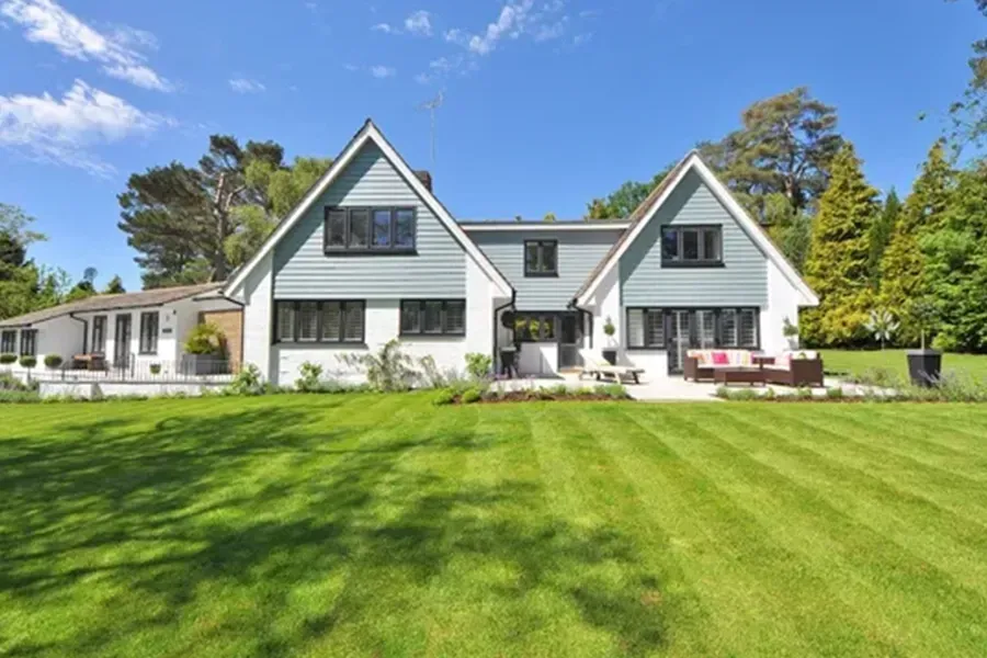 White farmhouse with dark trim on a sunny lawn, surrounded by trees and a blue sky.
