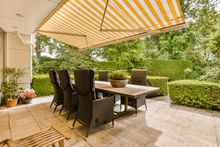 Patio with a table and chairs shaded by an awning; lush greenery in the background.
