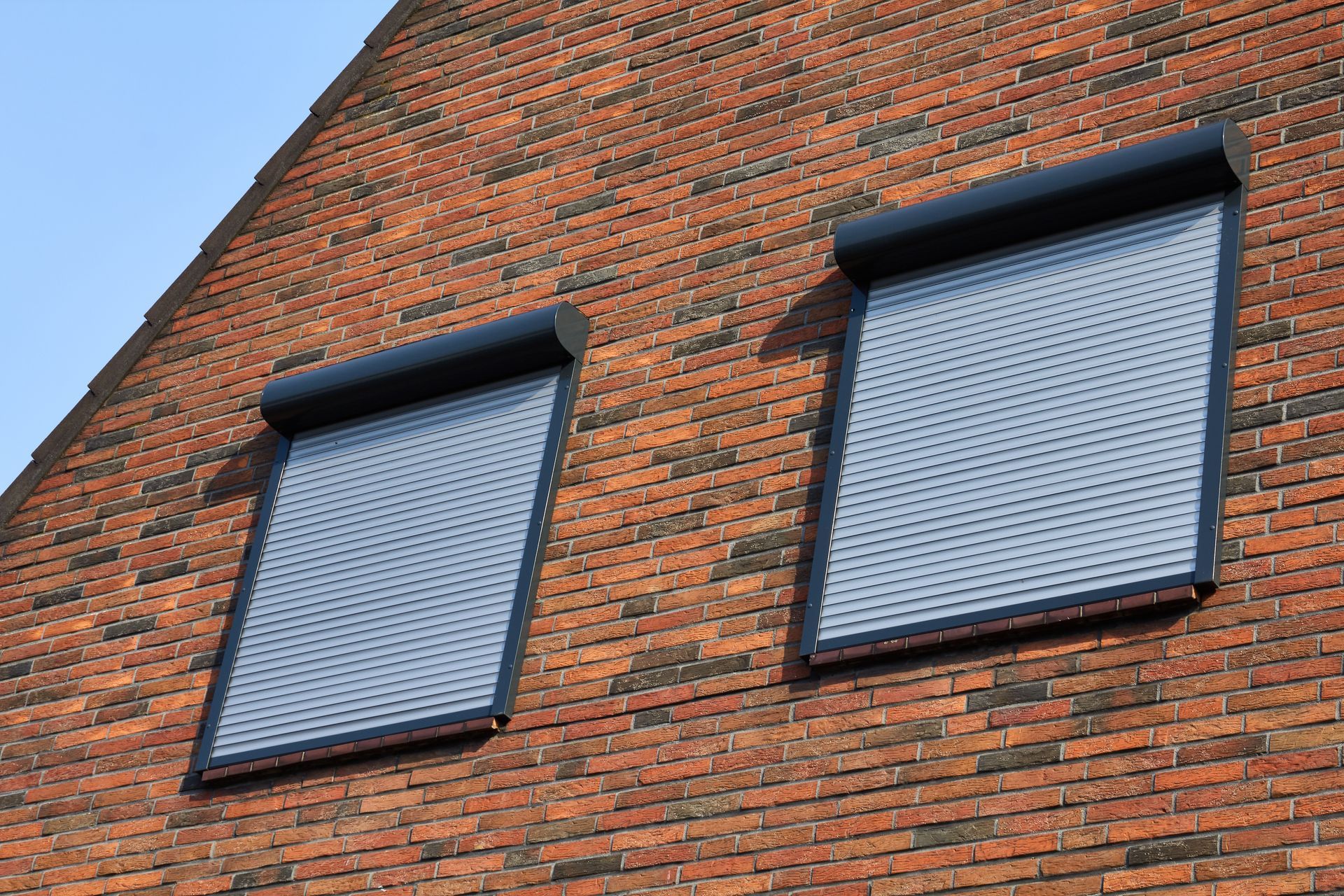 Two windows with closed, dark roller shutters on a red brick building.