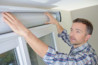 Man installing a white roller shutter above a window, indoors.