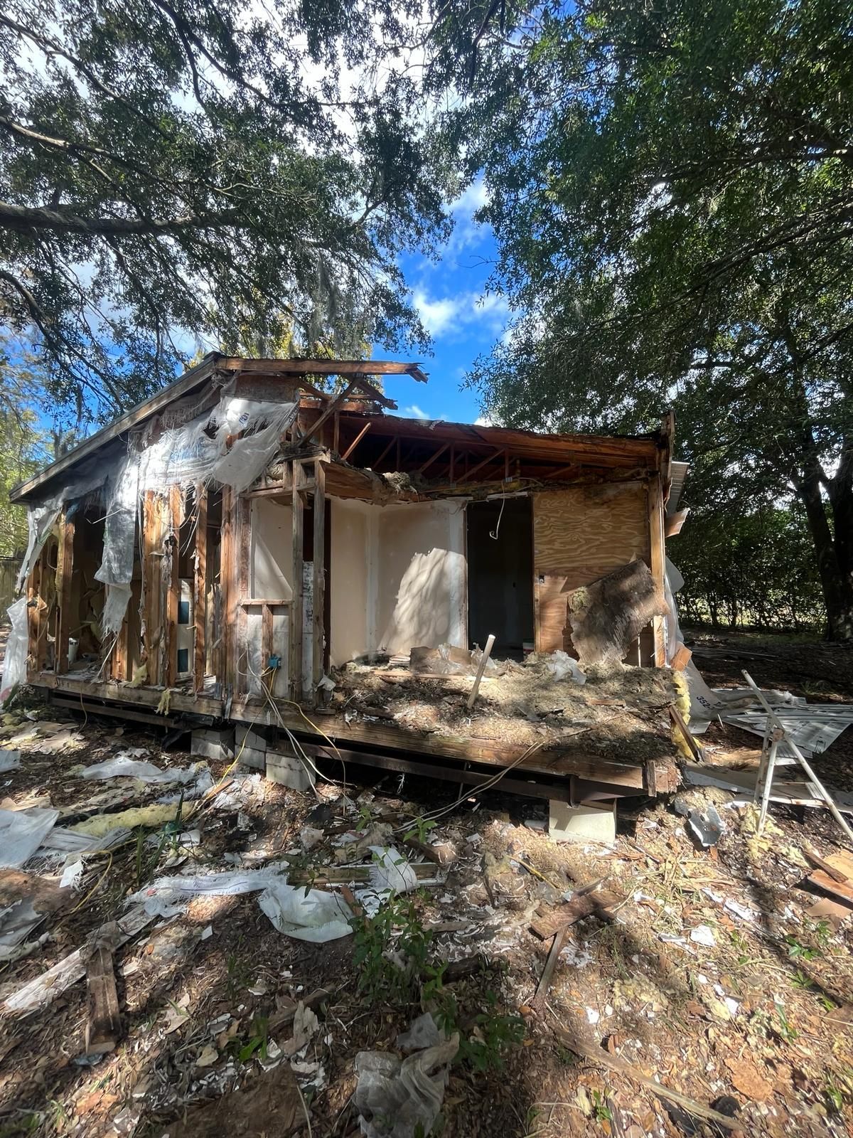 Exterior view of a dilapidated, partially demolished wooden building with exposed walls, under a partially visible blue sky.