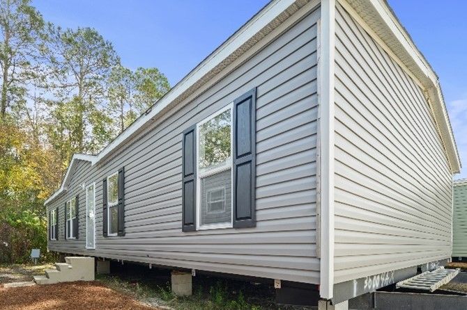 A gray, single-story manufactured home with white trim, black shutters, and a blue sky background.