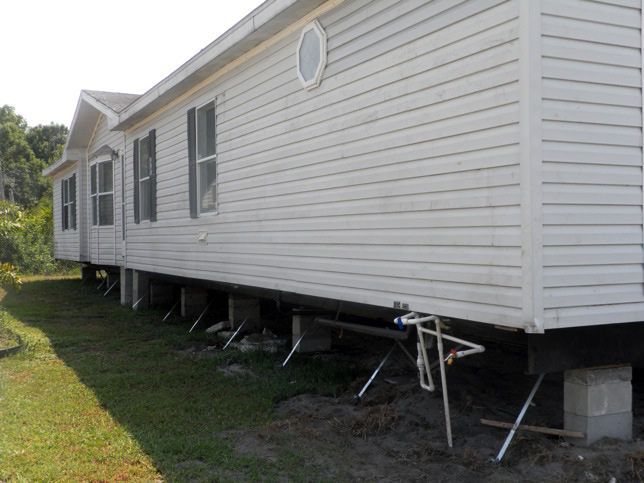 White mobile home on cinder blocks, with diagonal support beams in a grassy yard.