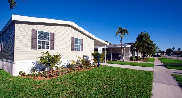 Tan single-story home with brown shutters and carport, lush green lawn, and clear blue sky. Palm trees and a sidewalk are also visible.