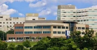Modern multi-story hospital building with tan and white facades, blue windows, and trees in front under a cloudy sky