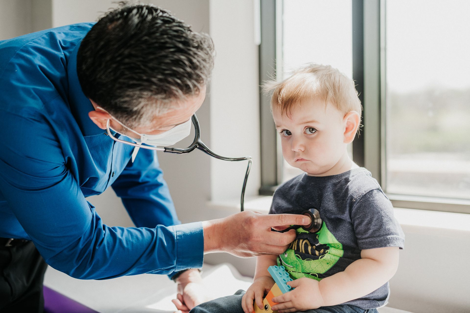 Doctor examining a young child with a stethoscope near a window