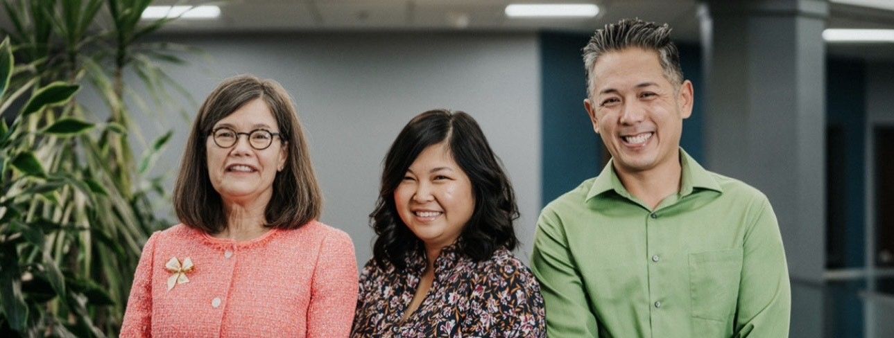Three coworkers smiling in an office, with plants and a blurred hallway behind them.